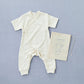 cream white cotton baby grow with a cross over front and a popper with poppers used to join the crotch area at the legs. with short sleeves and legs to the ankles. next to a draw string bag with the fog linen logo on it wit a linen illustration of the jumpsuit