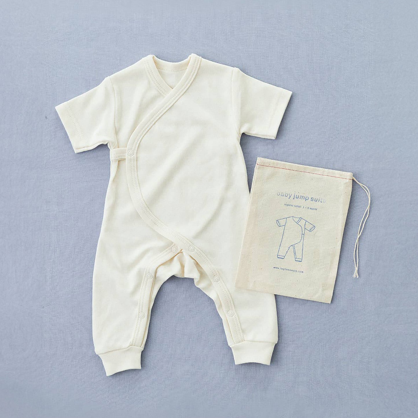 cream white cotton baby grow with a cross over front and a popper with poppers used to join the crotch area at the legs. with short sleeves and legs to the ankles. next to a draw string bag with the fog linen logo on it wit a linen illustration of the jumpsuit