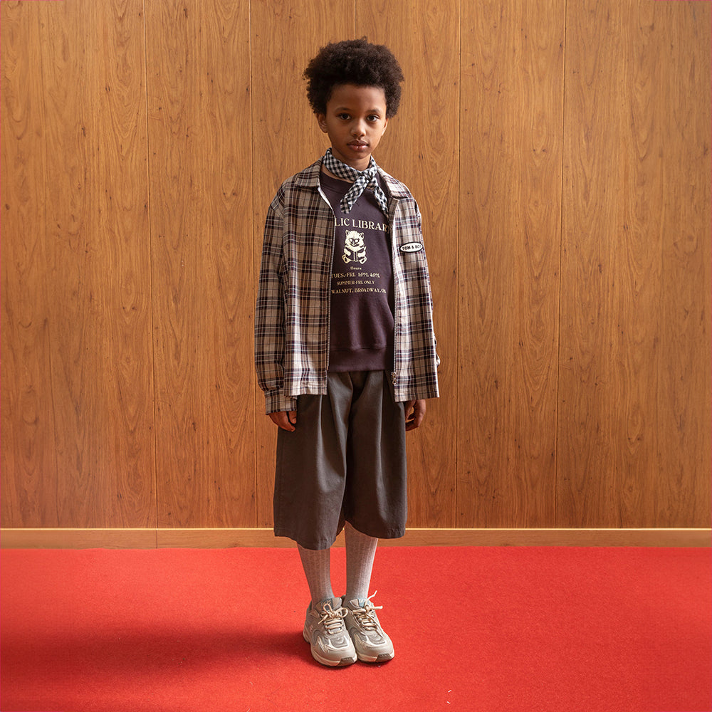 A mixed race boy stands in front of a brown panelled wall and a room with red flooring wearing a brown checkered over-shirt that says Tom&Boy to the right of the zip, inside of a white circle that has black bored Under the shirt is a brown Tom&Boy shirt that says 'public library' with the image of a bear reading a book. This is paired with grey wide leg shorts which fall below the knee. The boy is also wearing grey knee high socks with grey trainers and a checkered bandana around his neck. 