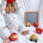 Baby in pajamas with apple pattern holding an apple, surrounded by apples and a toy car on a white background.
