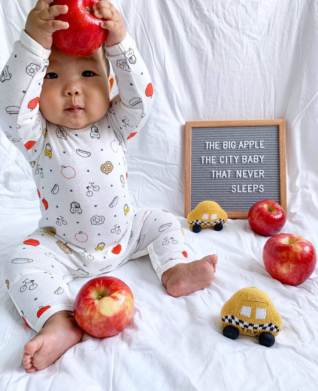 Baby in pajamas with apple pattern holding an apple, surrounded by apples and a toy car on a white background.