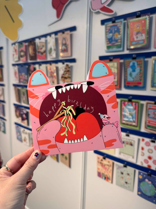 Person holding a pink cat-shaped birthday card with a colorful background of more cards.