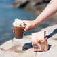Hand holding a Choc Shake with whipped cream by a scenic coastal background