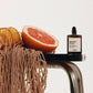 Bottle of Aromachologie perfume on a metallic stand with a grapefruit and a woven bag on a light background