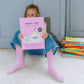 Child holding a pink book titled 'Kawaii Shop' in front of a stack of books on a light-colored floor.