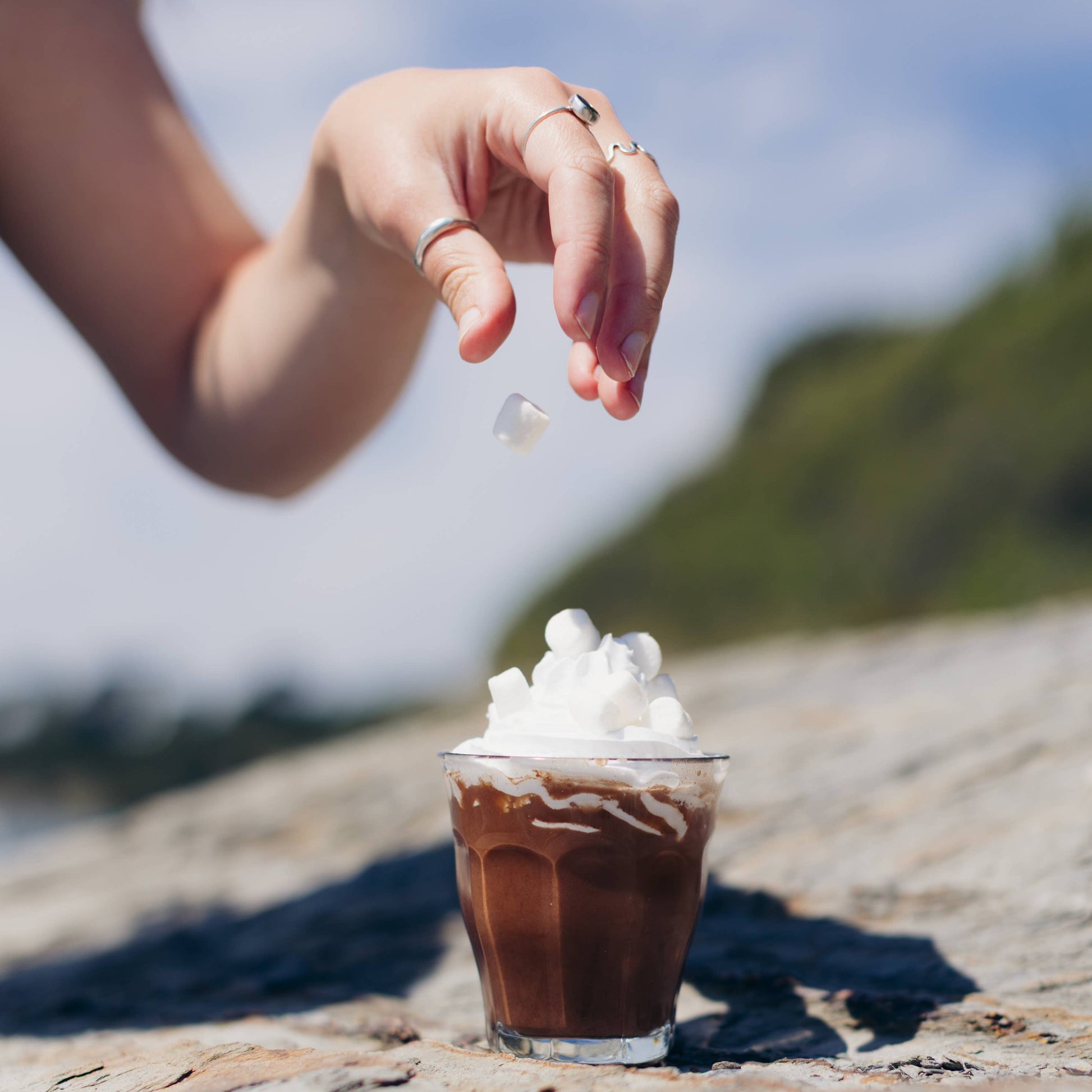 Hand sprinkling  mini marshmallows onto a small glass of chocolate milkshake with whipped cream on a stone surface.