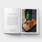 Open book showing a page with text and a photograph of food and a bottle on a table.