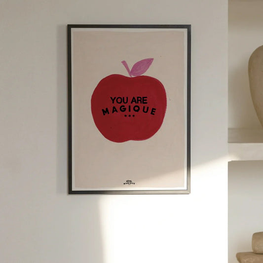 An image depicting a Hotel Magique Apple A3 print. The apple is red in colour with pink stem and leaf on top. The background is beige. “You are Magique” is written in black. The print is framed hung on a white wall.