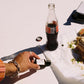 Person holding a bottle of Coca-Cola next to a bottle cap on a white surface.