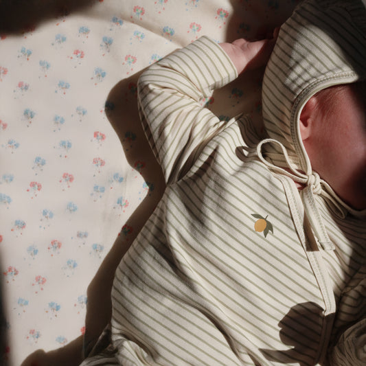 Close-up of a newborn wearing Konges Sløjd striped organic cotton clothing, featuring a delicate embroidered lemon detail. The baby is dressed in a cosy hooded outfit with tie fastening, lying on a soft floral-patterned blanket.