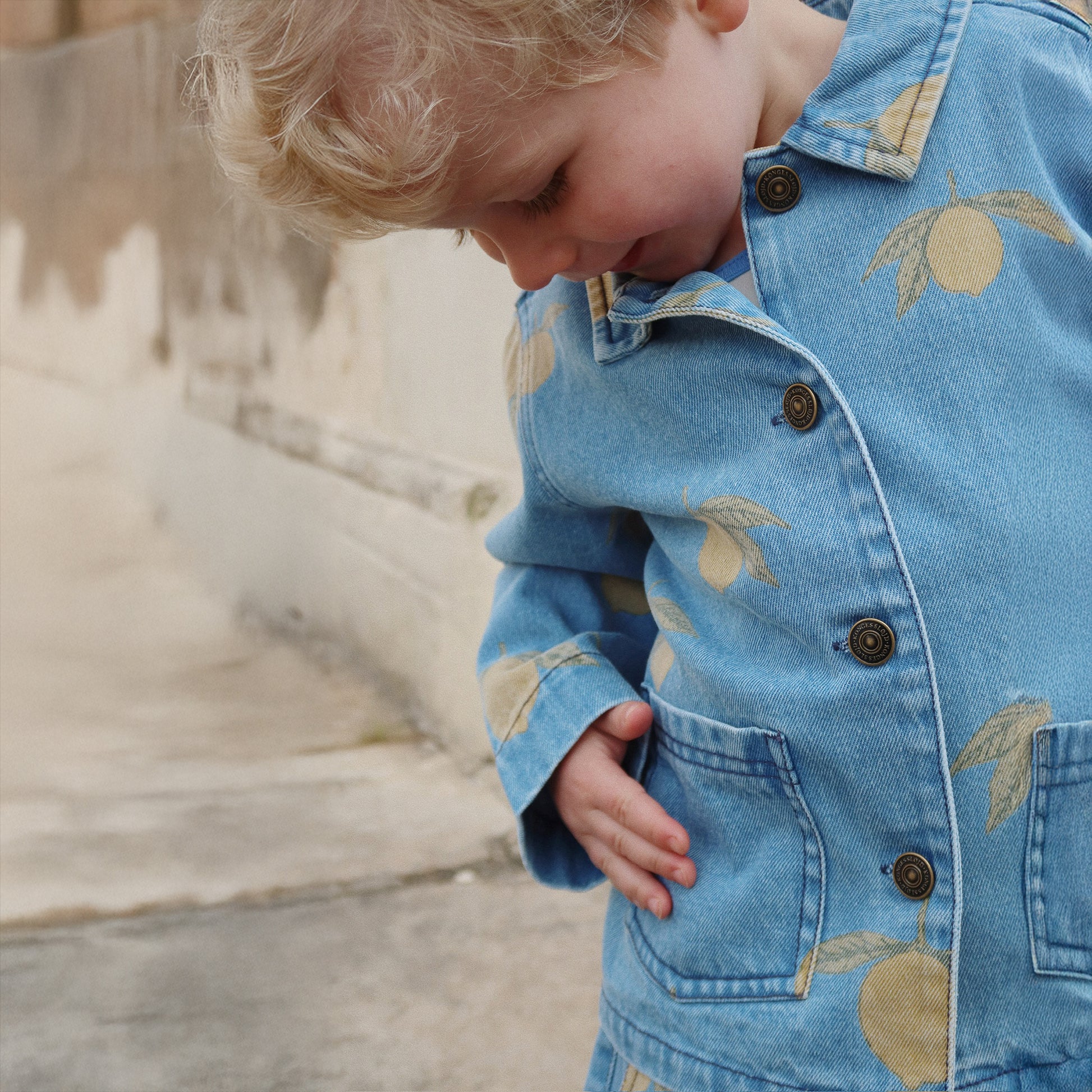  young child wearing the Konges Sløjd Magot Jacket in Lemon Denim. The image highlights the soft denim fabric, brass snap buttons, and delicate lemon print.