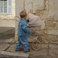 A young child in the Konges Sløjd Lemon Denim jacket and trousers pushing a vintage-inspired cream-coloured toy pram with pink polka dots, set against a rustic stone wall.