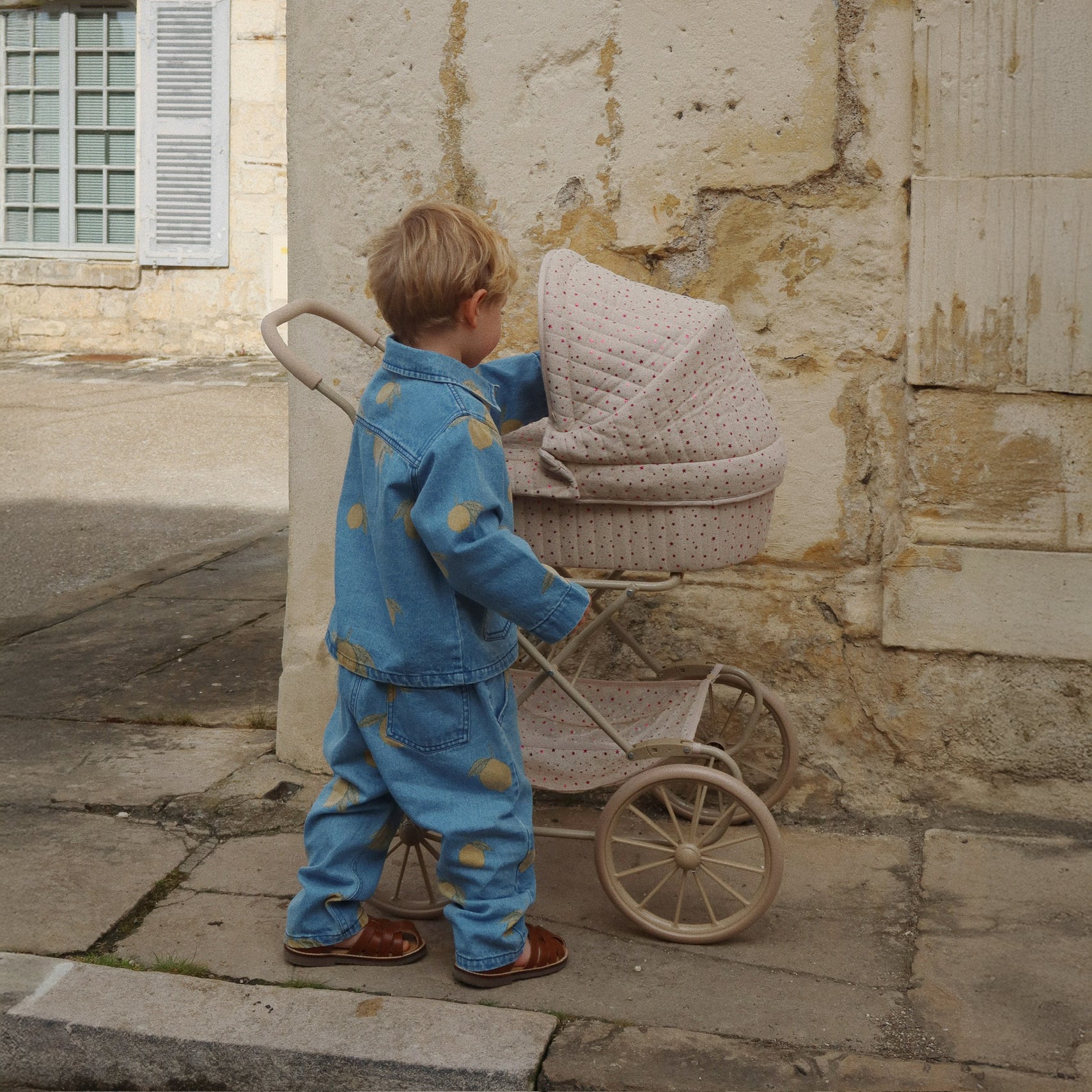 A young child in the Konges Sløjd Lemon Denim jacket and trousers pushing a vintage-inspired cream-coloured toy pram with pink polka dots, set against a rustic stone wall.