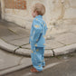 A young child wearing the Konges Sløjd Magot Jacket and matching Lemon Denim trousers, standing by a textured stone wall. The relaxed fit and playful lemon print are visible.