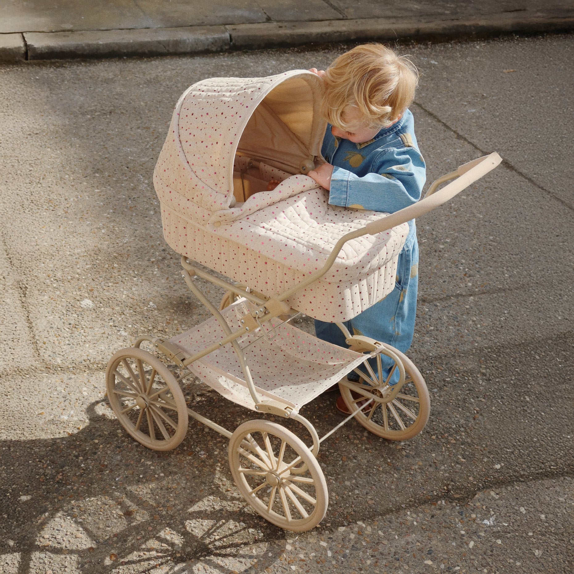 A top-down shot of a young child peeking into a vintage-style toy pram while wearing the Konges Sløjd Lemon Denim set. Sunlight highlights the soft quilted texture of the pram hood.