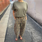 Model wearing Le Bon Shoppe Leopard Arc Pants with an olive green rolled-sleeve T-shirt and black flip-flops, standing in a sunlit urban alleyway.