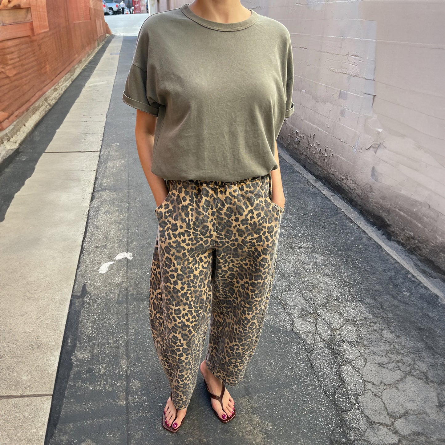 Model wearing Le Bon Shoppe Leopard Arc Pants with an olive green rolled-sleeve T-shirt and black flip-flops, standing in a sunlit urban alleyway.