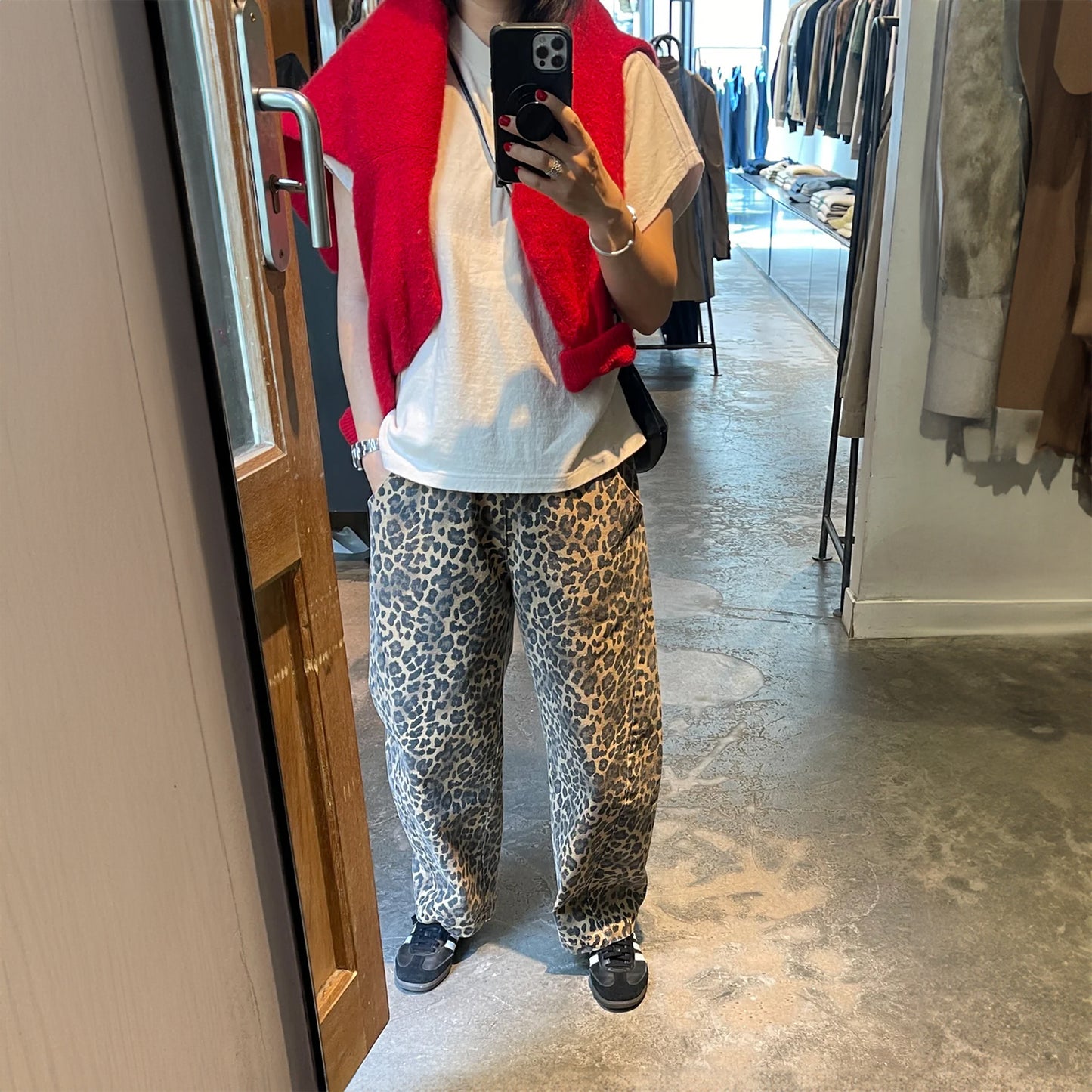 Person taking a mirror selfie wearing Le Bon Shoppe Leopard Arc Pants with a white T-shirt, bright red knit draped over the shoulders, and black trainers inside a modern boutique.