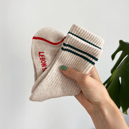 A hand holding two pairs of Le Bon Shoppe Boyfriend Socks in neutral tones, one with red details and the other with green stripes.
