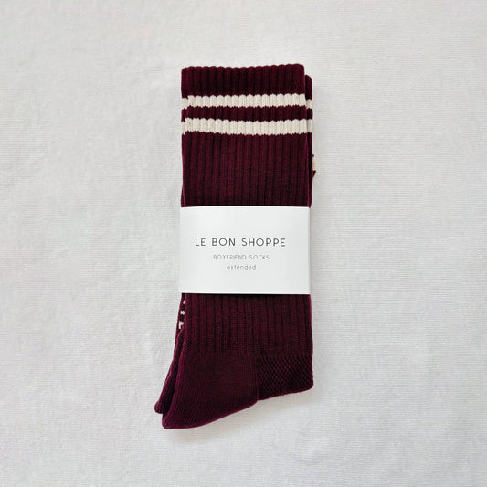 A folded pair of maroon Le Bon Shoppe Extended Boyfriend Socks with white stripes at the top, neatly wrapped in branded packaging, displayed on a clean white background.