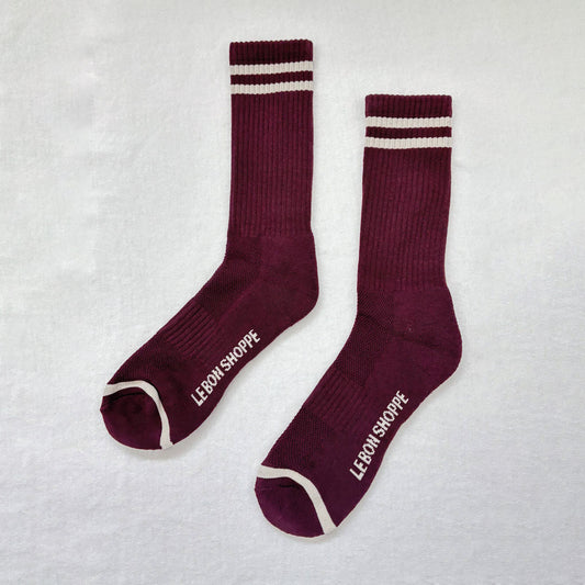 A pair of maroon Le Bon Shoppe Extended Boyfriend Socks laid flat, featuring ribbed texture, white contrast stripes, and branding on the sole, against a plain white background.