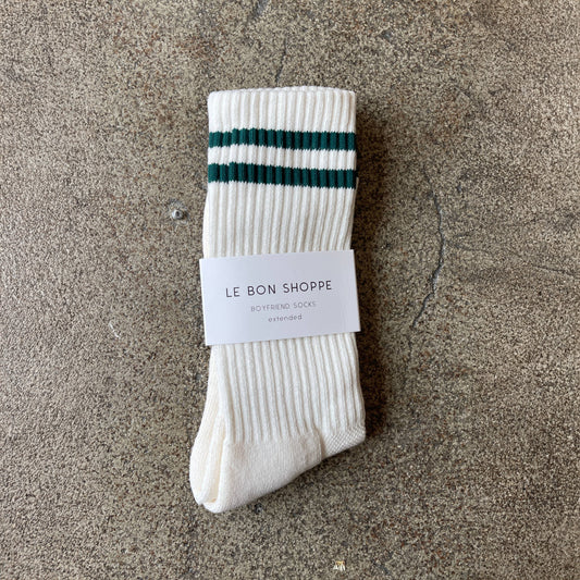 A folded pair of parchment white Le Bon Shoppe Boyfriend Socks with dark green stripes, wrapped in branded packaging, laid on a concrete surface.