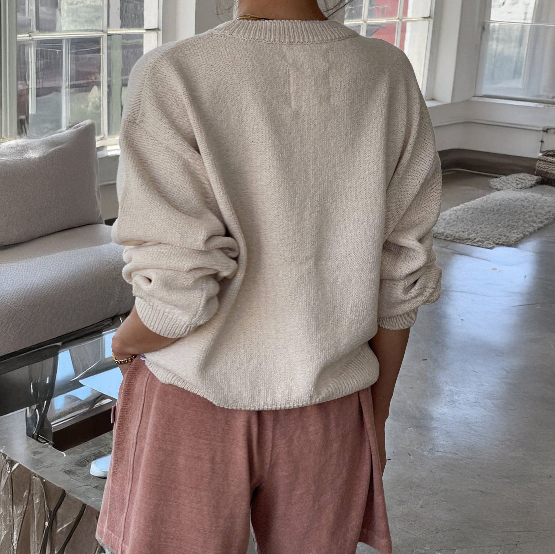 Person wearing a naturel sweater and pink pants in a room with a couch and windows.