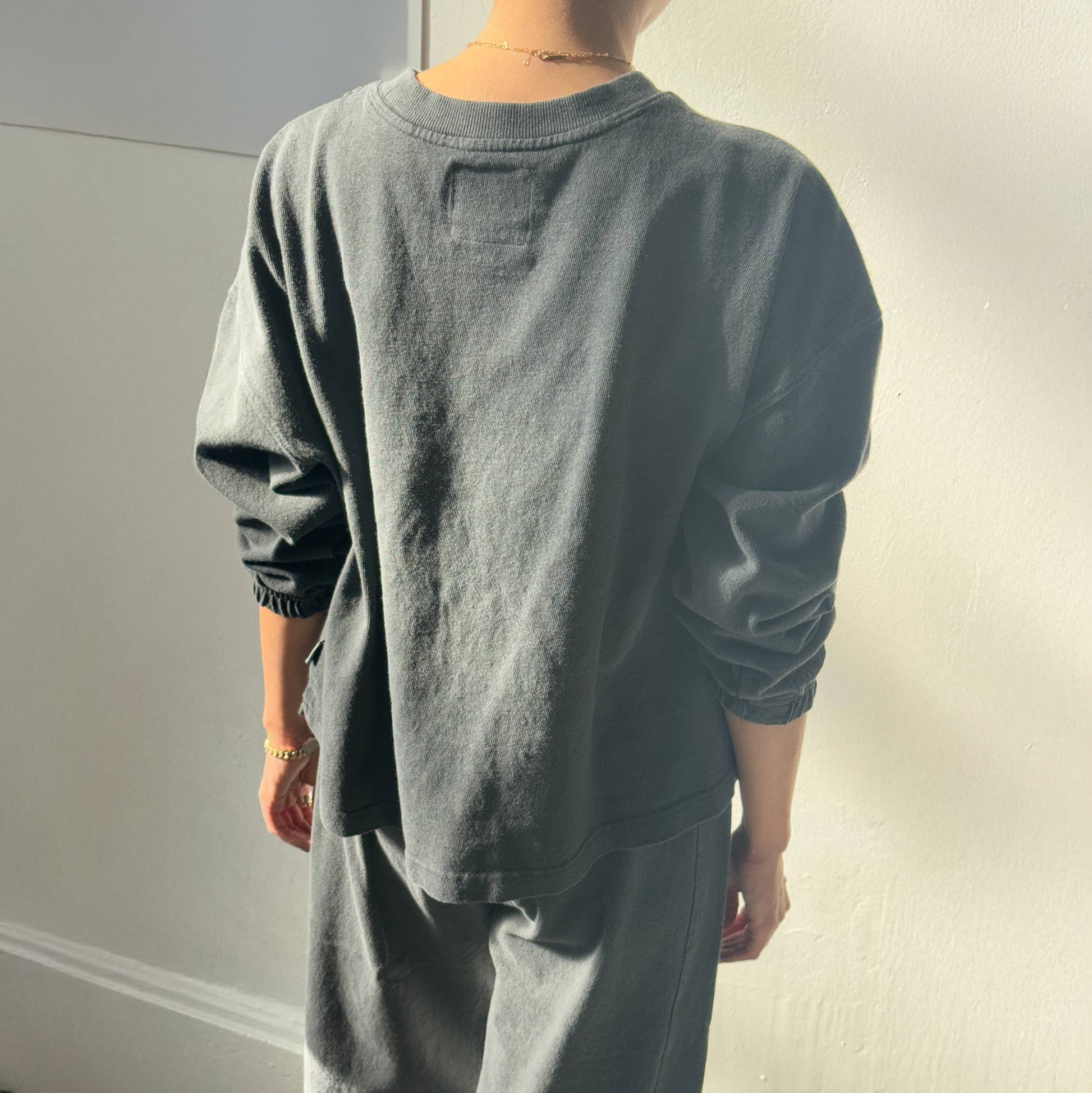 Back view of the Le Bon Shoppe Naturelle Tee in Coal – A relaxed-fit, heavyweight cotton long-sleeve tee with a dropped shoulder and ruched sleeve detail, worn by a model in soft lighting.