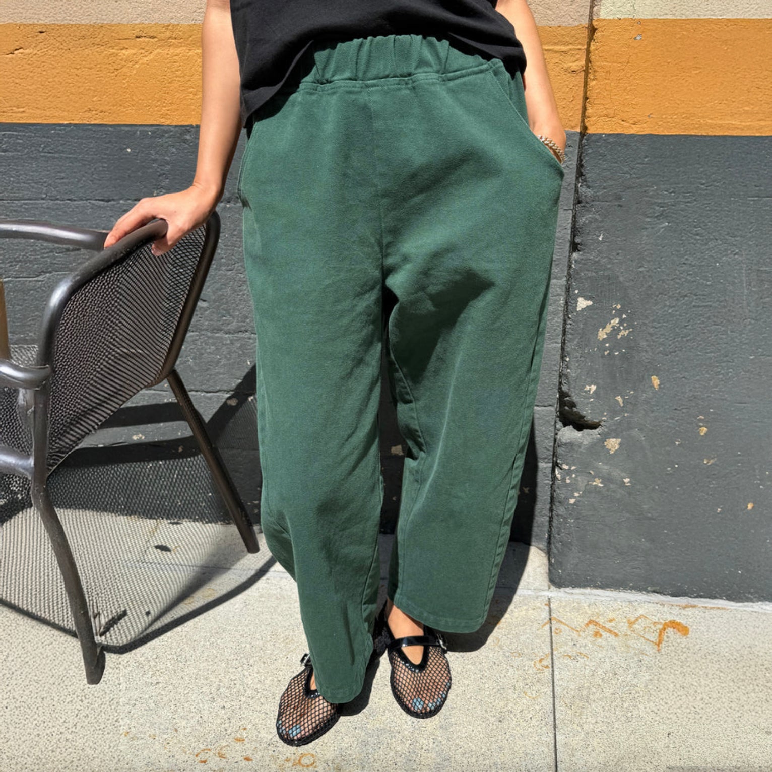 Person wearing green pants standing on a sidewalk next to a chair.
