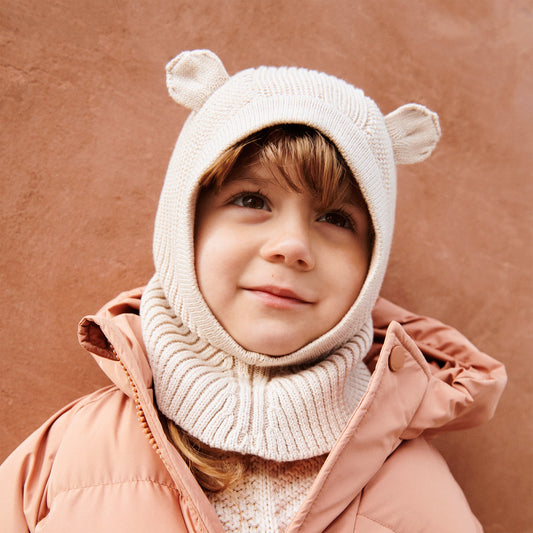 Child wearing a pink coat and bear-themed hat against a brown background