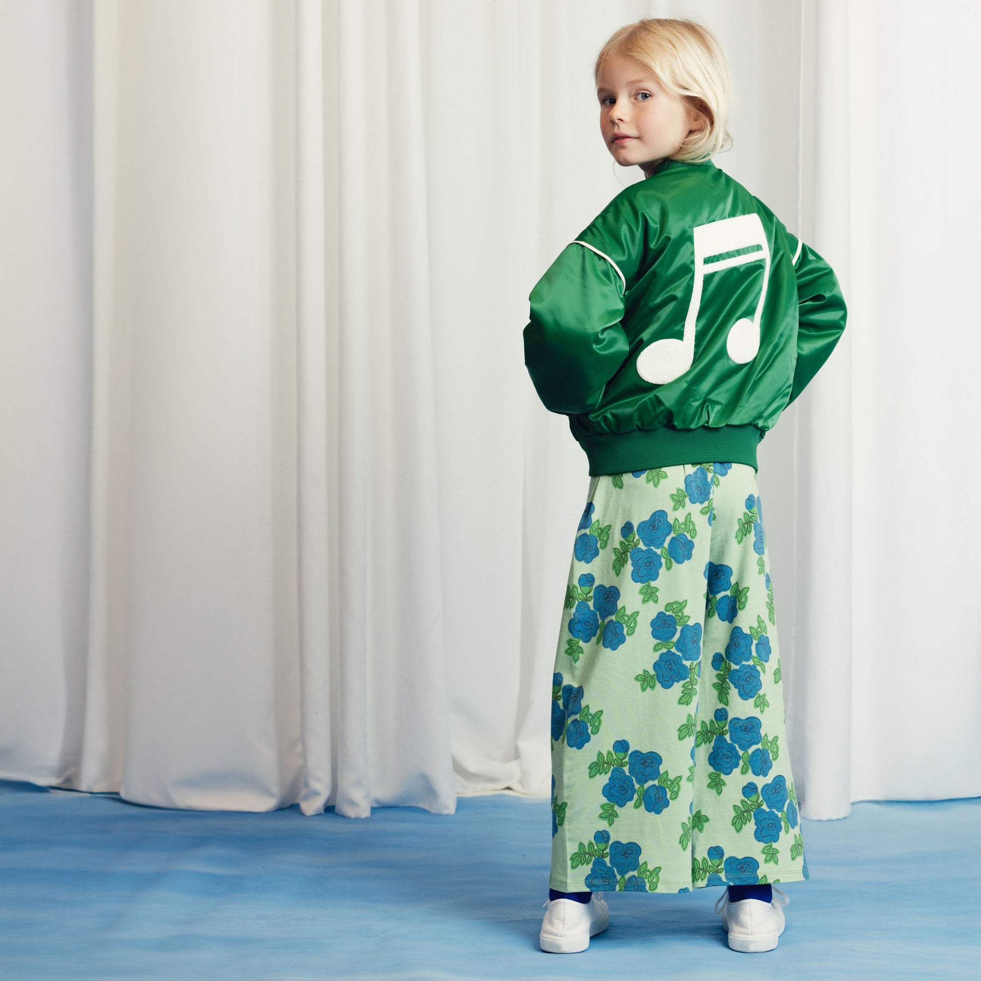 A young child wearing a green satin bomber jacket with an oversized white embroidered music note on the back, paired with wide-leg trousers in a blue floral print.