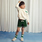 Young boy wearing a cream Mini Rodini sweatshirt with green piping details and a small embroidered chameleon motif on the chest. Paired with green knit shorts, chameleon-print socks, and white sneakers. He stands with one hand in his hair, looking off to the side.