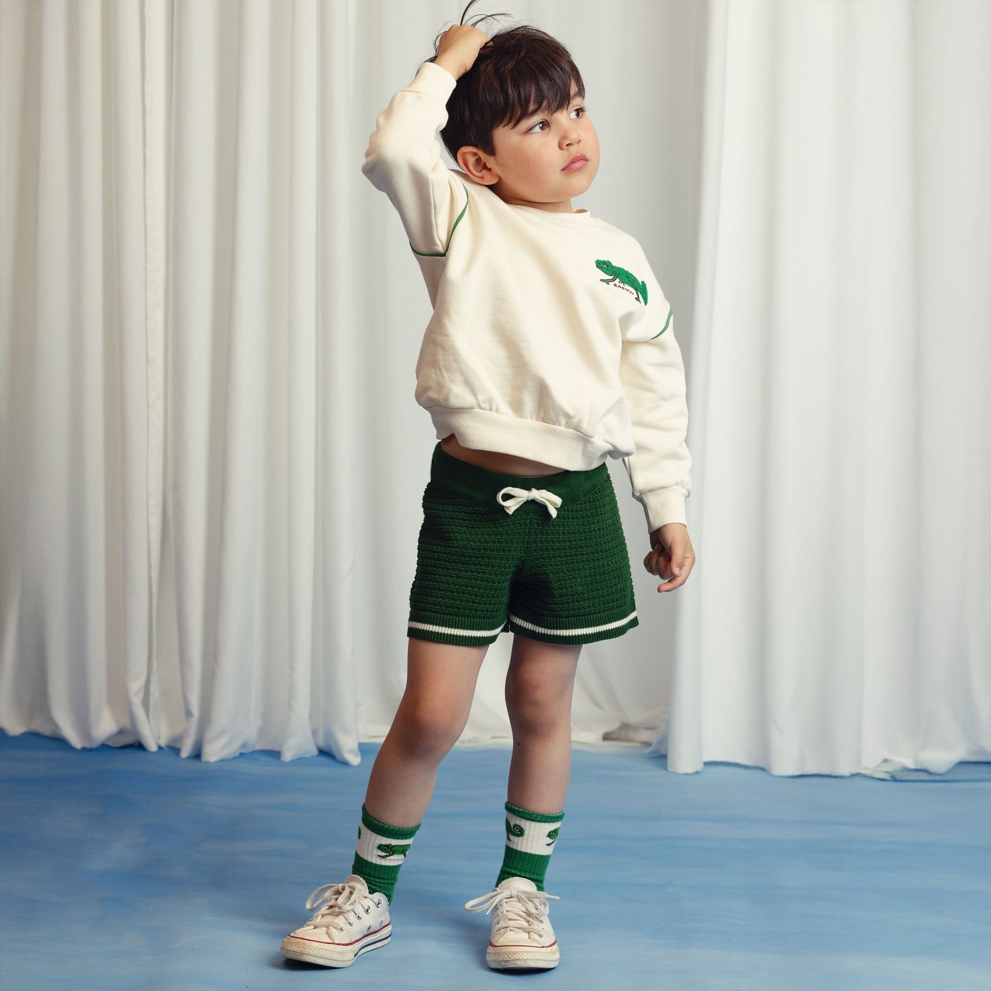 Young boy wearing a cream Mini Rodini sweatshirt with green piping details and a small embroidered chameleon motif on the chest. Paired with green knit shorts, chameleon-print socks, and white sneakers. He stands with one hand in his hair, looking off to the side.