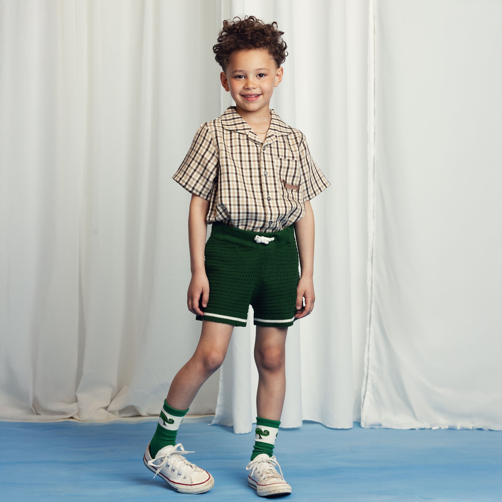 Young boy wearing a beige and brown checkered short-sleeve shirt paired with textured green knit shorts featuring a white drawstring. Styled with green and white chameleon-print socks and white sneakers. He smiles at the camera against a cream curtain backdrop.