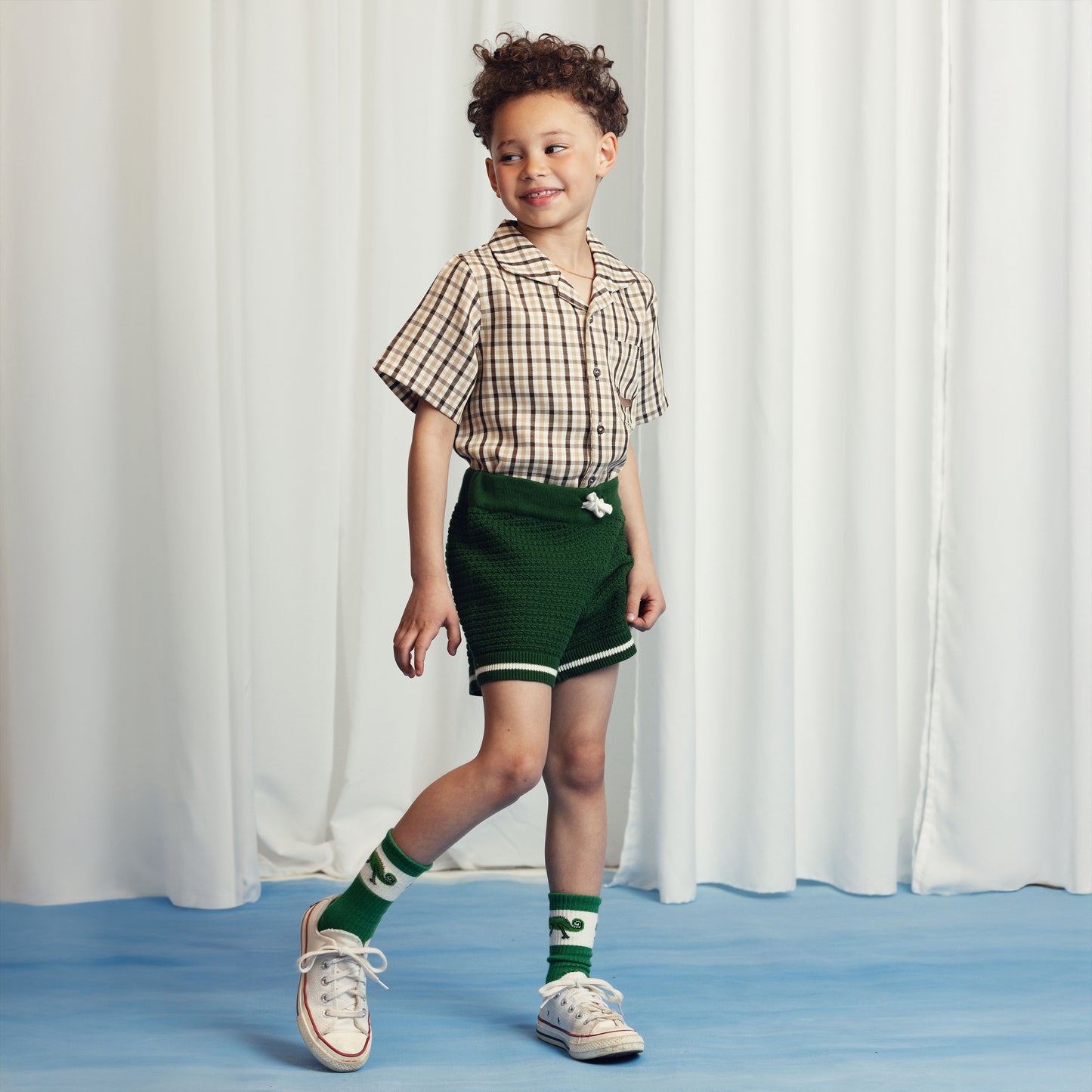 Young boy wearing a checkered short-sleeve shirt and green knit shorts, mid-step with a cheerful expression. He pairs the outfit with green chameleon-print socks and white sneakers.