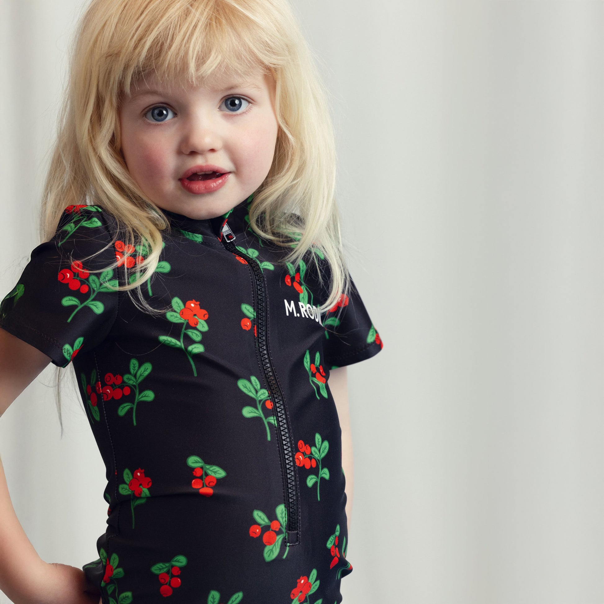 A young child wearing a black short-sleeve zip-up swimsuit featuring an all-over red berry print and the Mini Rodini logo on the chest.