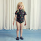 A child standing in Mini Rodini’s berry-print swimsuit paired with navy blue jelly sandals, smiling at the camera.