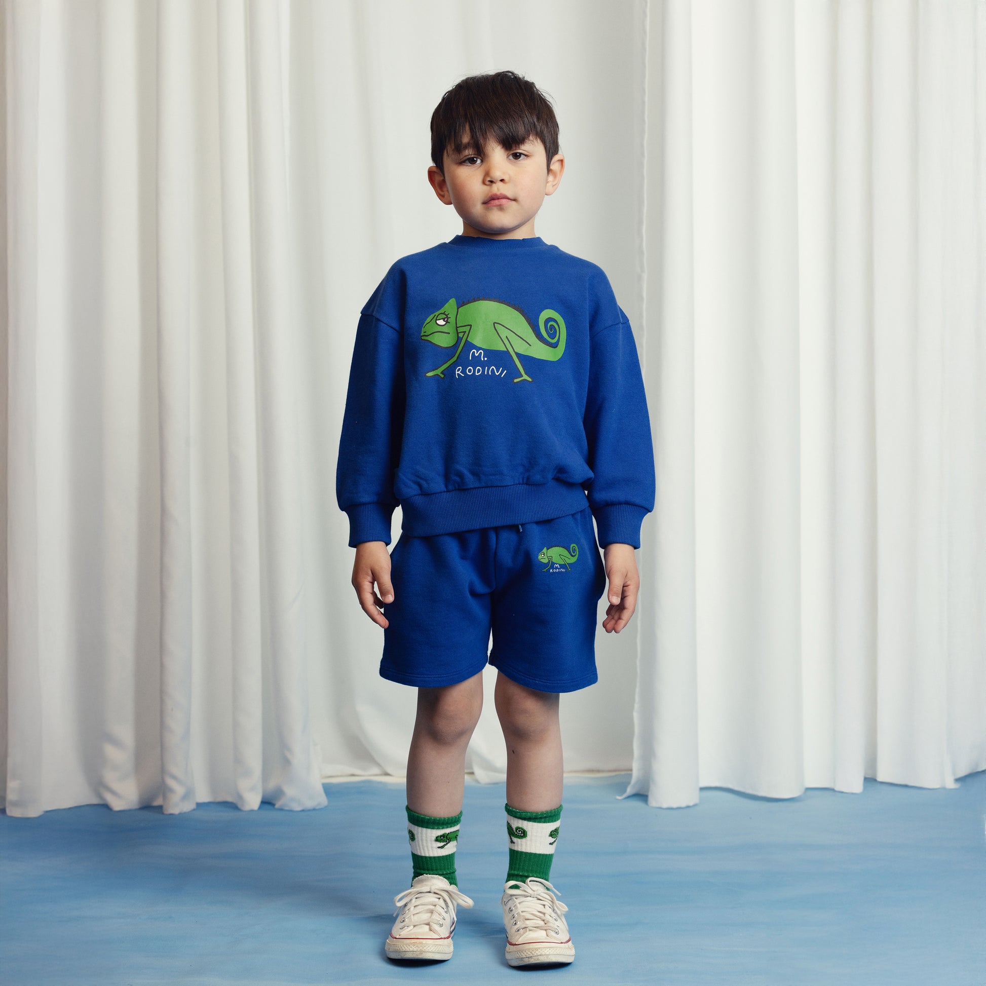 Young boy wearing a royal blue Mini Rodini sweatshirt featuring a bold green chameleon graphic with "M. Rodini" text. Styled with matching blue shorts embroidered with a small chameleon motif, green and white striped socks, and white sneakers. He stands against a neutral cream curtain backdrop with a calm expression.