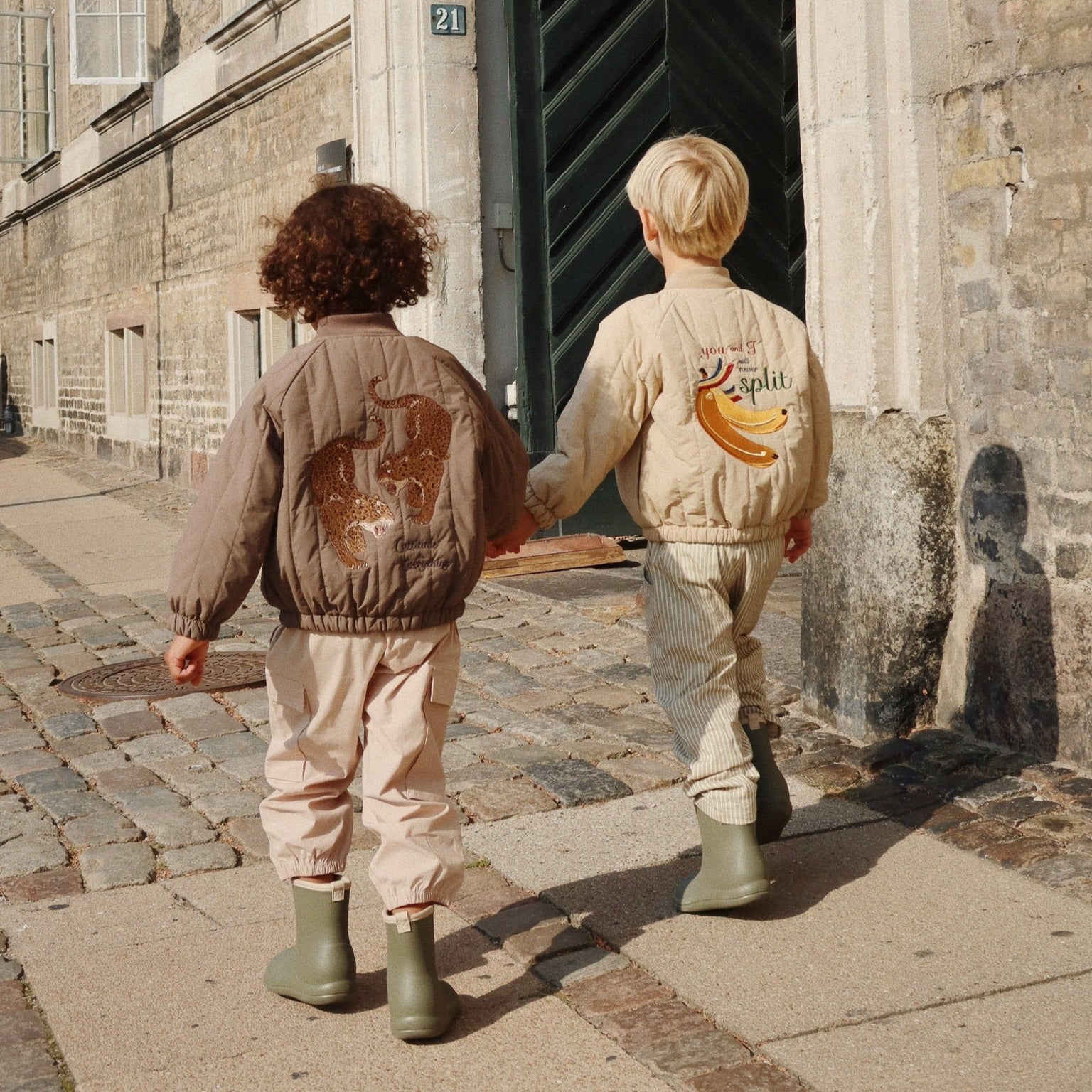 Two children walking hand in hand on a cobblestone street, wearing matching outfits with graphic designs.