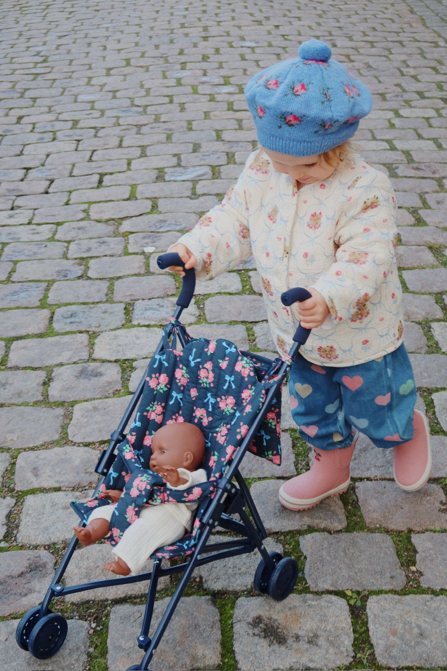 Child pushing a floral stroller with a doll on a cobblestone street