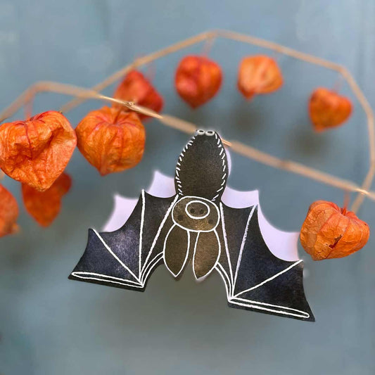 A die cut greeting cars in the shape of a bat hangs upside down on a dried physalis branch