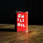 A red tin of Olea Pia chilli oil sits on a wooden table against a black background