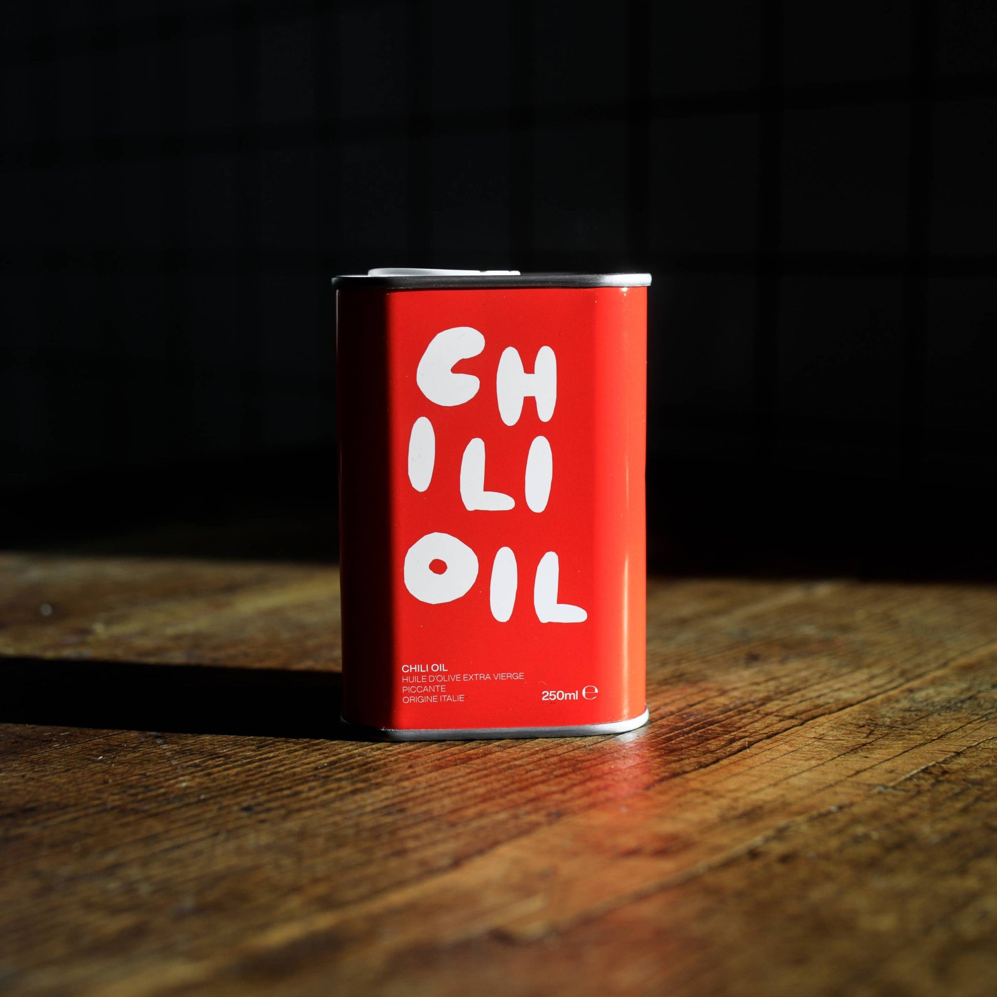 A red tin of Olea Pia chilli oil sits on a wooden table against a black background