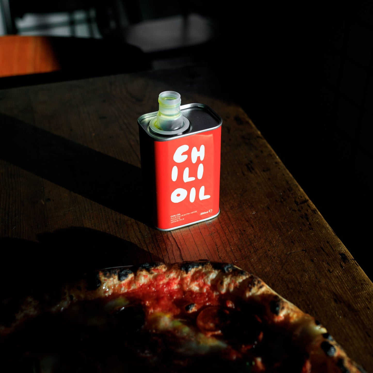 An open red tin of Olea Pia chilli oil sits on top of a wooden table in a dark room next to a freshly baked pizza