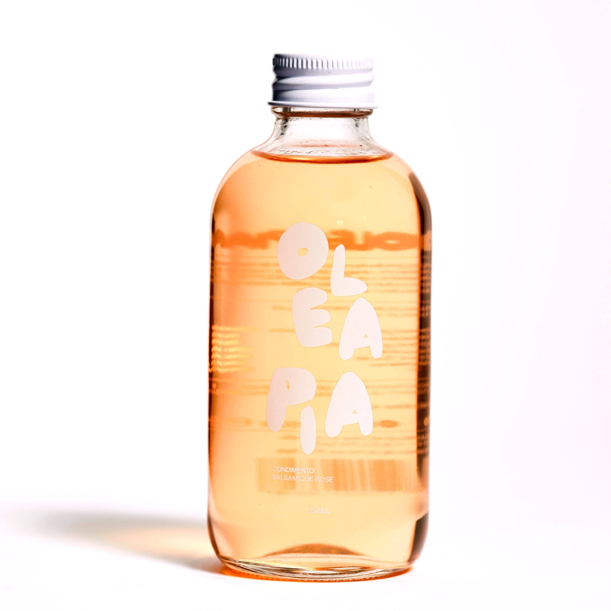 a glass bottle with a white screw top sits in front of a white background. The bottle has white writing on it that you can see from the other side but cannot read. On the front you can make out the Olea Pia logo in white. The bottle contains a pinkish liquid which is Olea Pia rosé balsamic vinegar