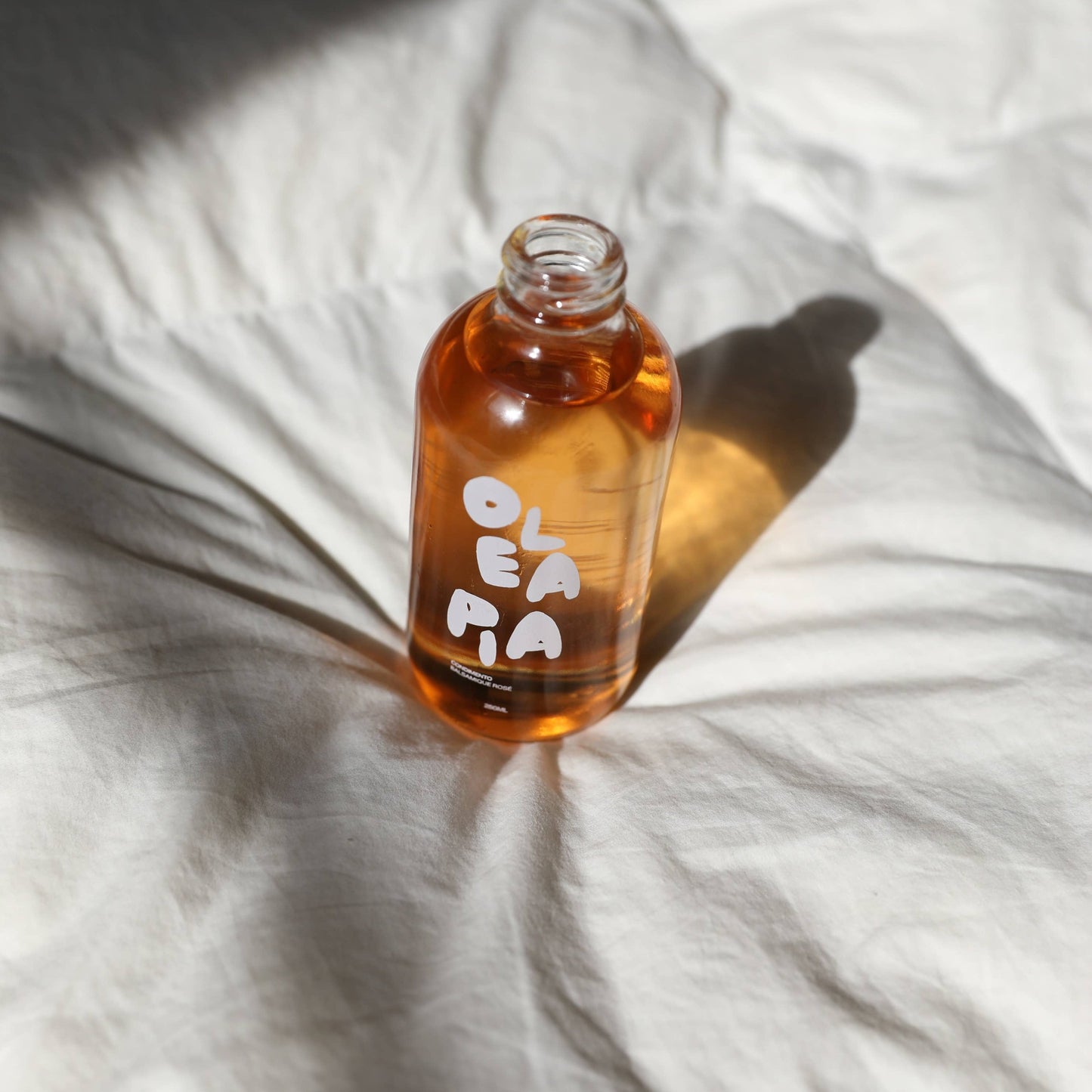 An open glass bottle sits on top of what looks like a white duvet. The bottle is open and contains a pinkish balsamic vinegar - the colour is the same a rosé wine. The bottle has OLEA PIA written on in in white text