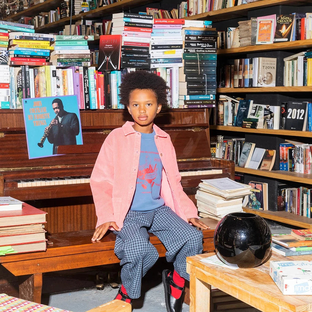 A mixed race boy sits on a bench in front of a piano in what looks like a bookshop of library, with books stacked high on every surface. A John Coltrane vinyl album sits behind him. He is wearing the Tom & Boy blue bagel sweatshirt underneath a pink overskirt, with checked trousers and red socks.