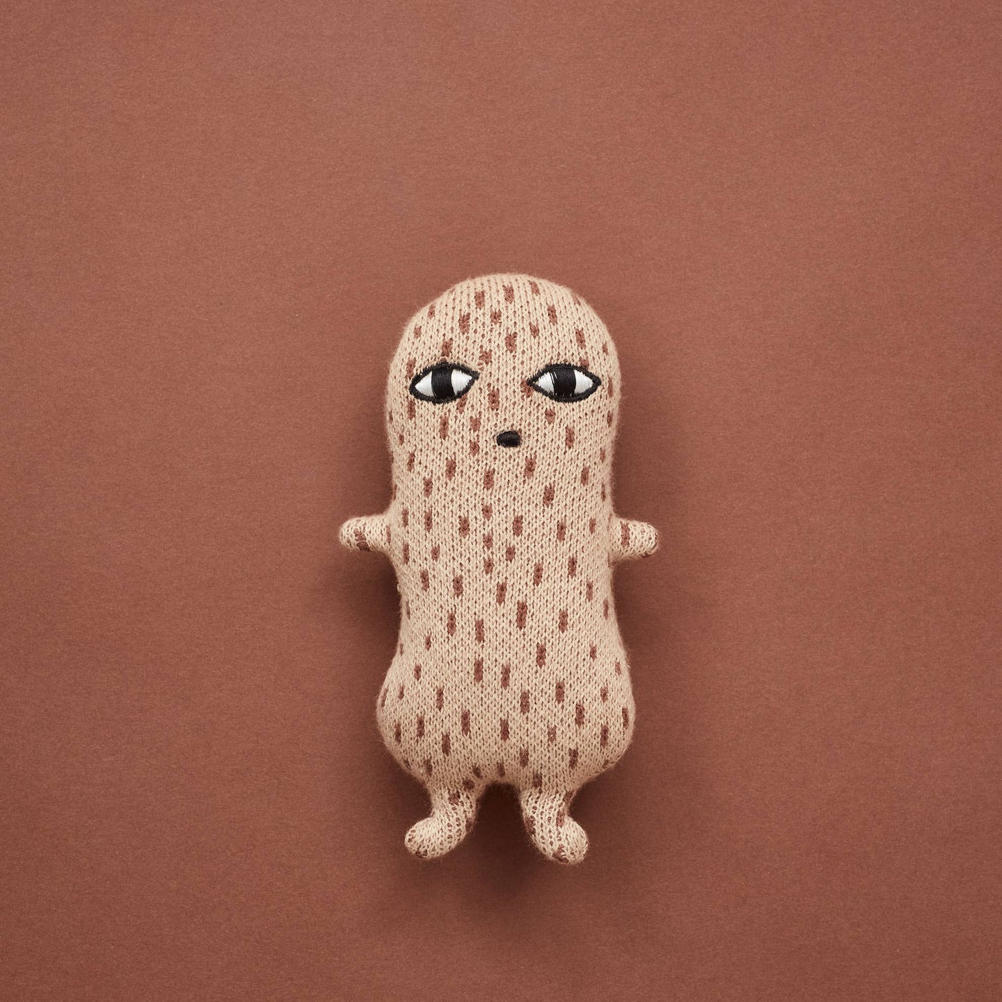 On a rust backdrop the peanut Donna Wilson naive illustrated style knitted rattle. A creamy brown with brown dashes all over with an embroidered little mouth and eyes, with two little feet and arms