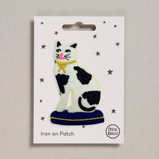 An embroidered patch depicting a black and white cat sitting on a blue cushion. It has a link nose and a gold bow around its neck. The backing card is white with black stars 