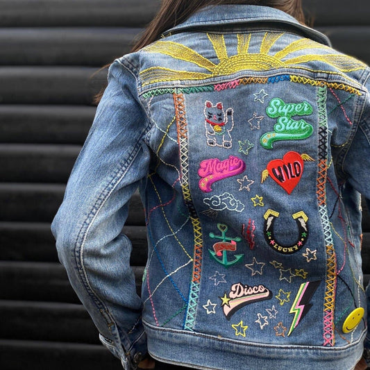 A medium-wash denim jacket covered in vibrant embroidered patches and hand-stitched details. The patches include playful words like "Super Star" and "Disco," a lucky horseshoe, a waving lucky cat, and lightning bolts, all arranged with bright crisscrossed stitching and a golden embroidered sunburst on the back yoke.